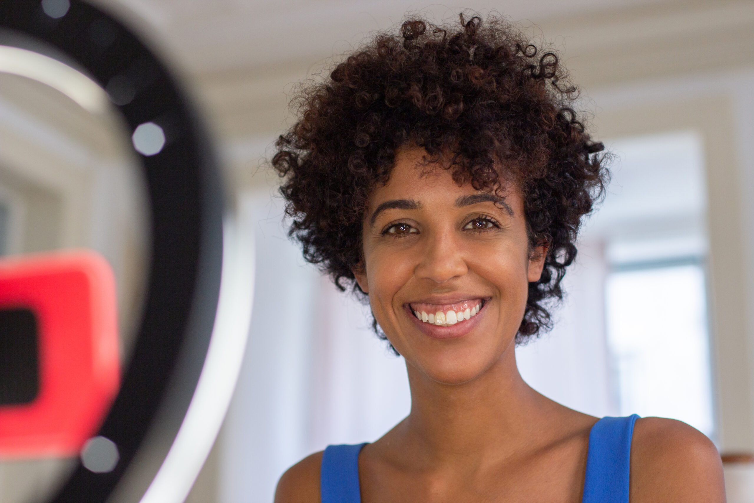 Close-up of happy female beauty blogger. Young Latin woman with brown eyes and dark curly hair smiling widely when vlogging indoors looking straight at camera.  Cosmetics, beauty, make up blogs concept